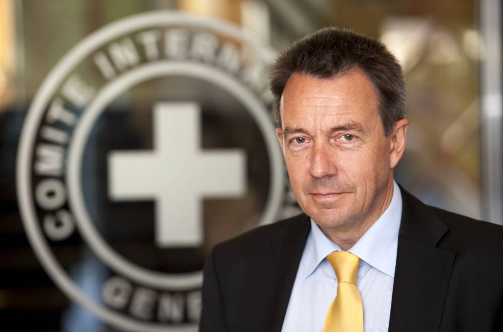 Photograph of the President of the International Committee of the Red Cross (ICRC), Peter Maurer. The ICRC President is wearing a black suit jacket with a white shirt and yellow tie. Behind him is a glass surface with the ICRC logo in white, the main element of which is a cross.